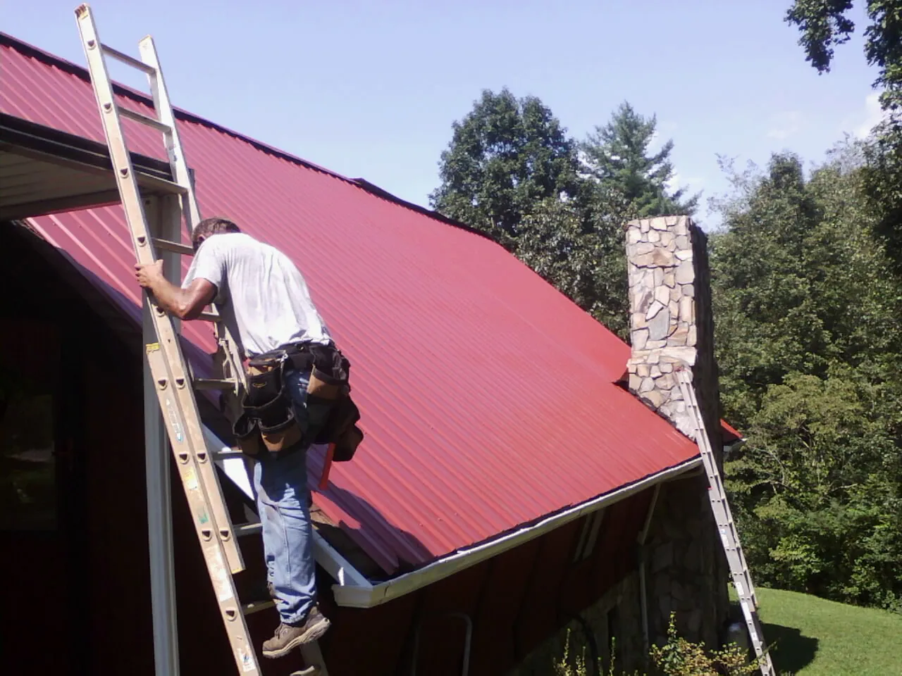 Roofer on a ladder beside a red metal roof and stone chimney — Soffit & Fascia Repair by Roofing Brothers in Lake Stevens